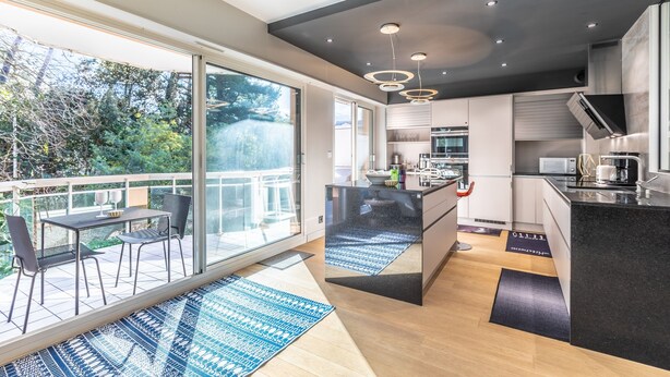 Een moderne keuken met een groot schiereiland, inbouwapparatuur en een schuifdeur naar een balkon met buitenmeubels.