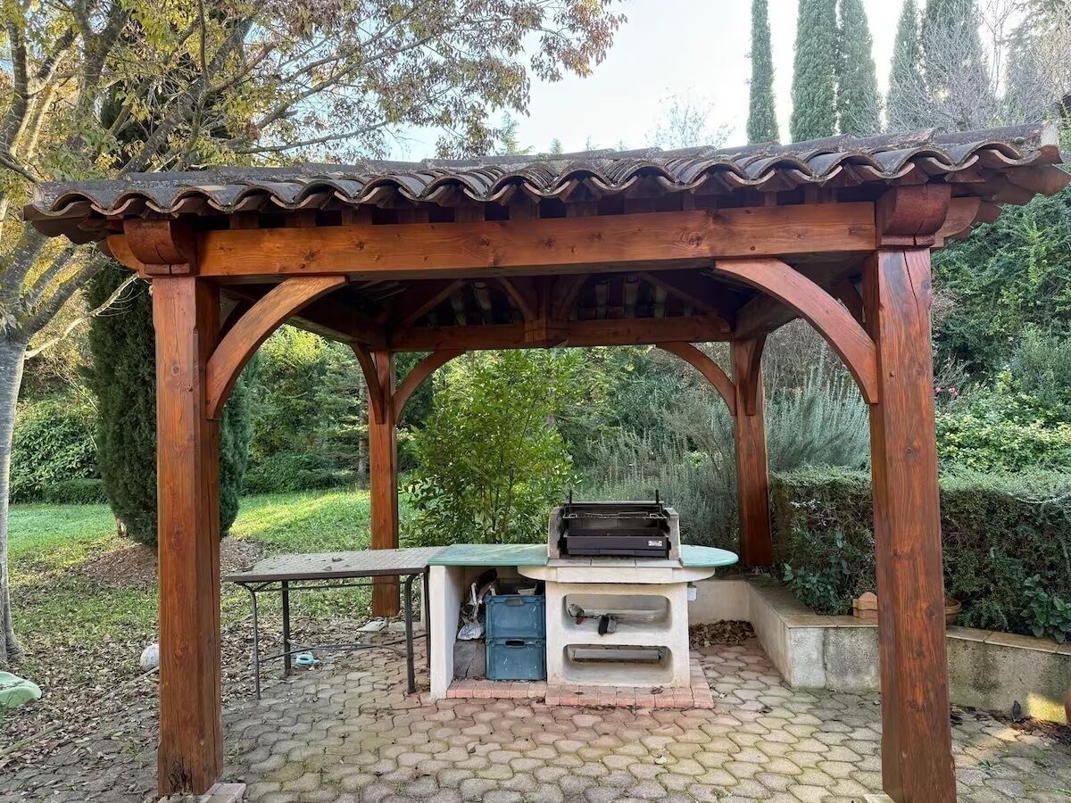 Een buitenbarbecuezone met een houten pergola, een barbecue en een tafel.