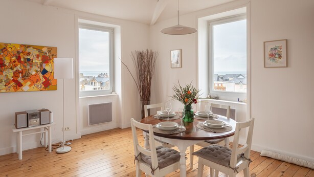 Spiseområde med et rundt bord, hvide stole, en vase med blomster og udsigt over byen gennem store vinduer.