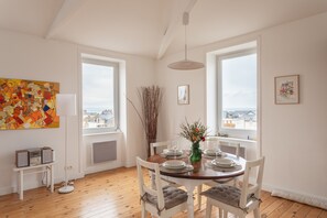 A dining area with a round table, white chairs, a vase with flowers, and a city view through large windows. - Au Fil de L'eau - 300 m de la plage (Pléneuf-Val-André)