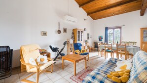 En rummelig stue med træloft, sofa, gyngestol og spiseområde med bord og stole.