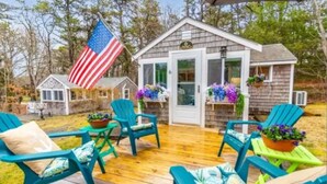 Une petite cabane en bois avec un porche, des fauteuils Adirondack verts et un drapeau américain suspendu.