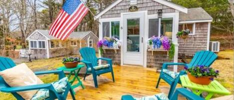 A small wooden cabin with a porch, green Adirondack chairs, and a hanging American flag.