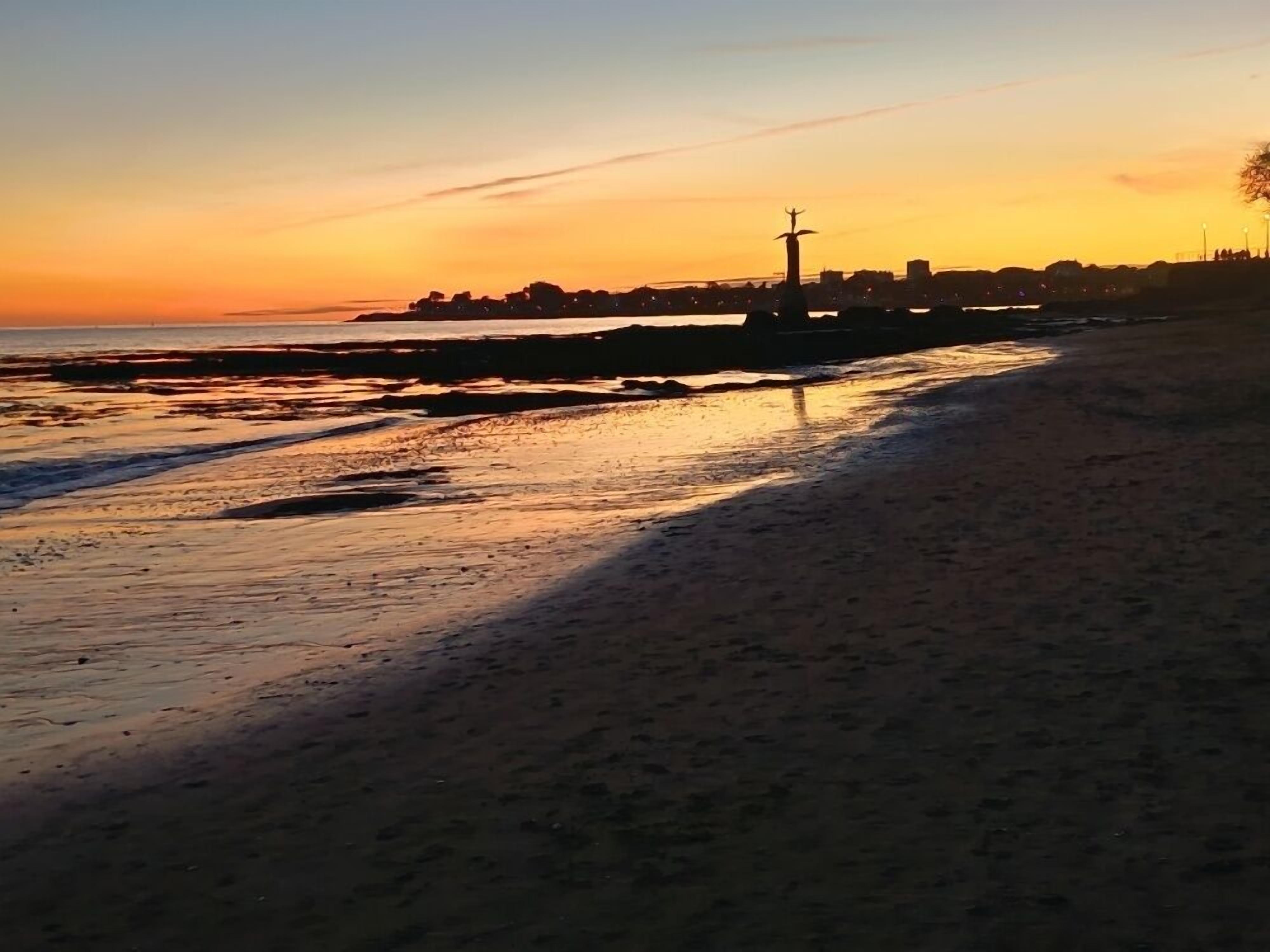 A beach at sunset with a pier and distant buildings.