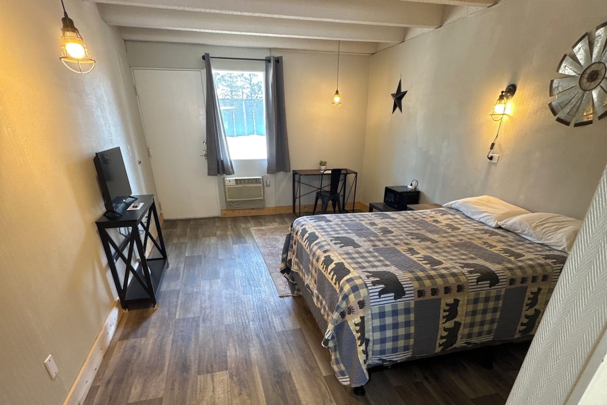 Un dormitorio con suelo de madera, una cama con colcha estampada, un escritorio con silla, una mesita con lámpara y una ventana con cortinas.