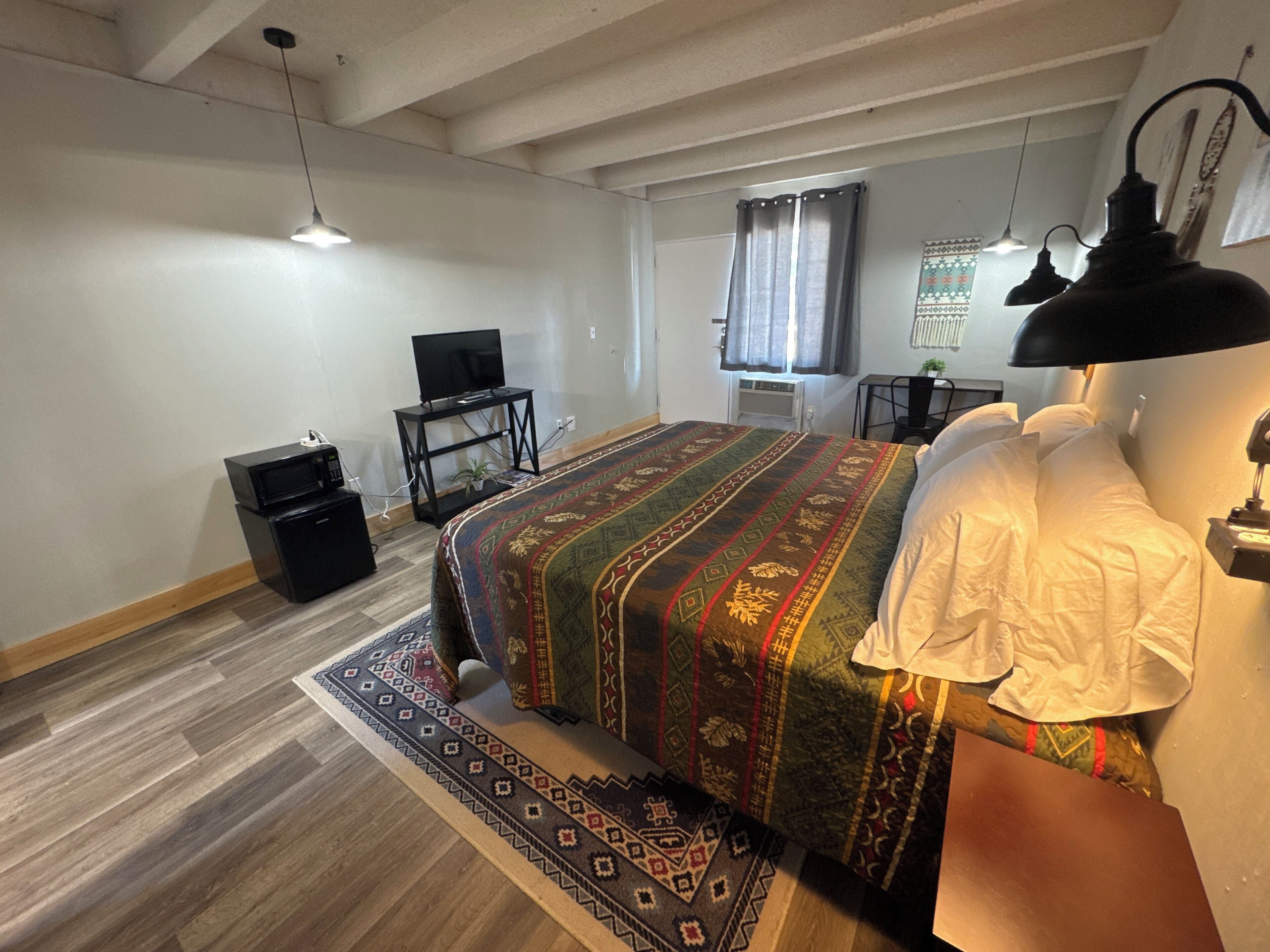 A bedroom with a large bed, a TV on a stand, a desk with a chair, and a radiator.