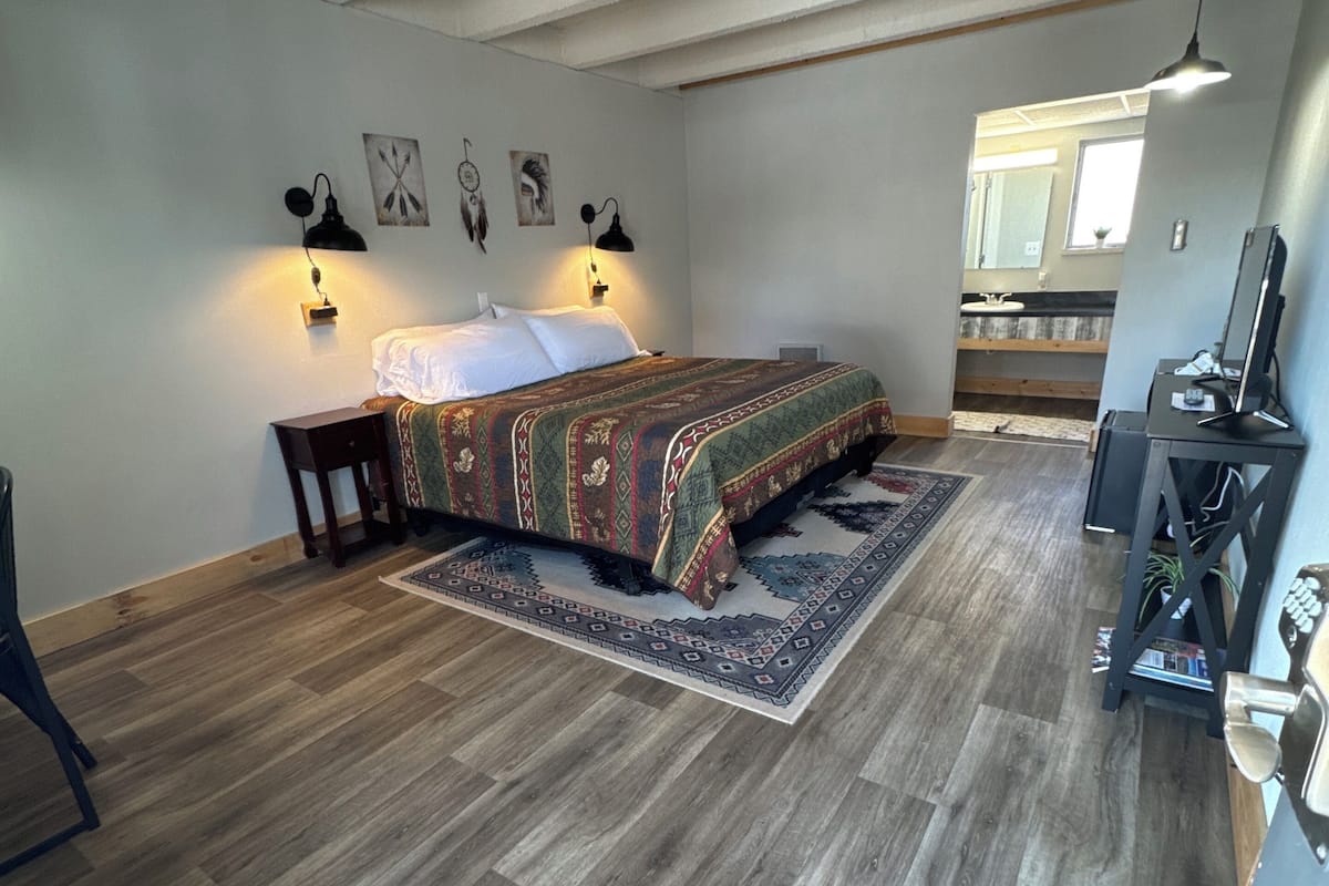 Un dormitorio con cama, suelo de madera, alfombra, escritorio con silla y espejo.