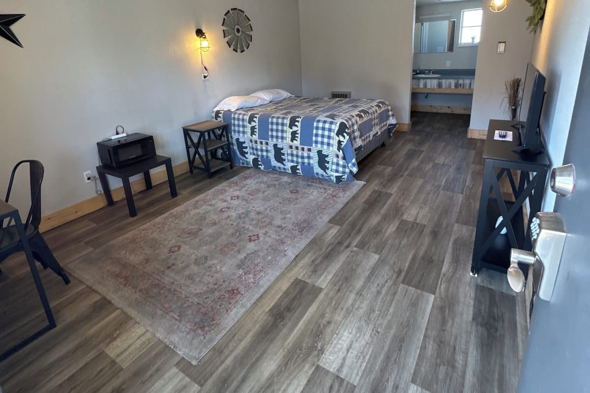 Una habitación compacta con cama, escritorio, silla, mesita pequeña, un reloj de pared y una lámpara colgante.