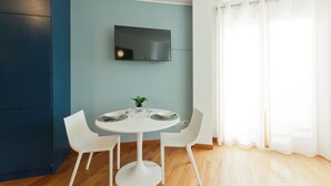 Un espace repas moderne avec une table ronde, des chaises blanches, un téléviseur à écran plat fixé au mur et une grande fenêtre ornée de rideaux légers.