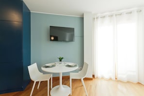 Ein moderner Essbereich mit einem runden Tisch, weißen Stühlen, einem flachbildfernseher an der Wand und einem großen Fenster mit transparenten Vorhängen.