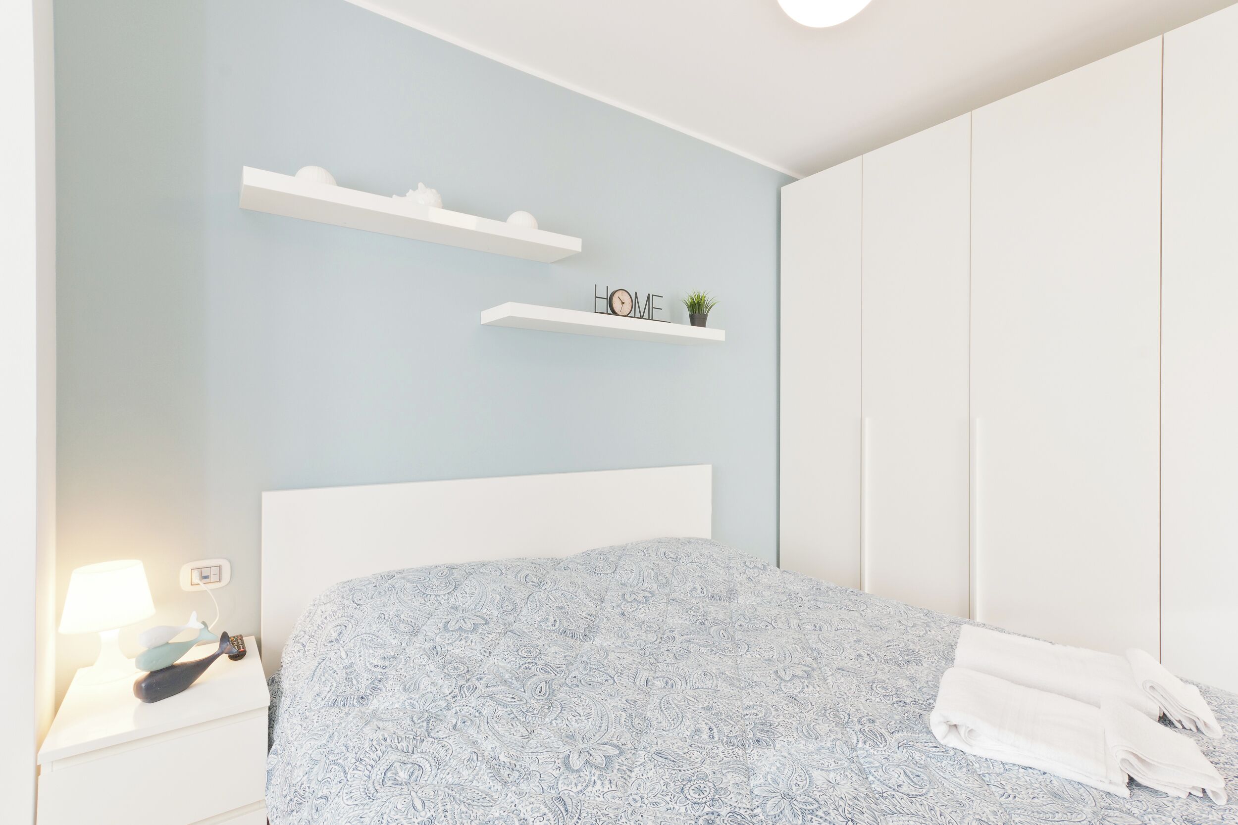 Une chambre à coucher comprenant un lit, une table de chevet, une lampe, une étagère murale avec une plante et une armoire.