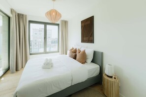 A modern hotel room with a large bed, a view through the window, and a wooden side table with a candle. - Large apartment with balcony, 3 BR and 2 Bathrooms (Antwerpen)