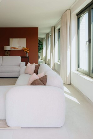 A modern living room with a large white sectional sofa, a small side table with a lamp, and a painting on the wall. - Large apartment with balcony, 3 BR and 2 Bathrooms (Antwerpen)