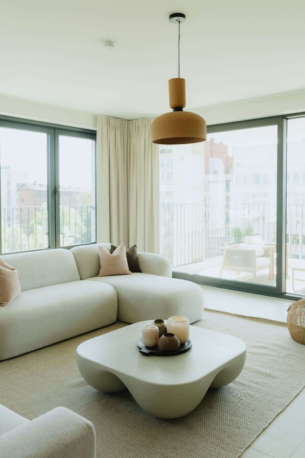 A modern living room with a white sofa, a round coffee table with decorative items, a hanging pendant light, and large windows with a view of buildings. - Large apartment with balcony, 3 BR and 2 Bathrooms (Antwerpen)