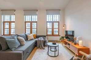 Una sala de estar moderna con un sofá gris, una mesa redonda de centro, un televisor sobre un mueble de madera y ventanas grandes con persianas.