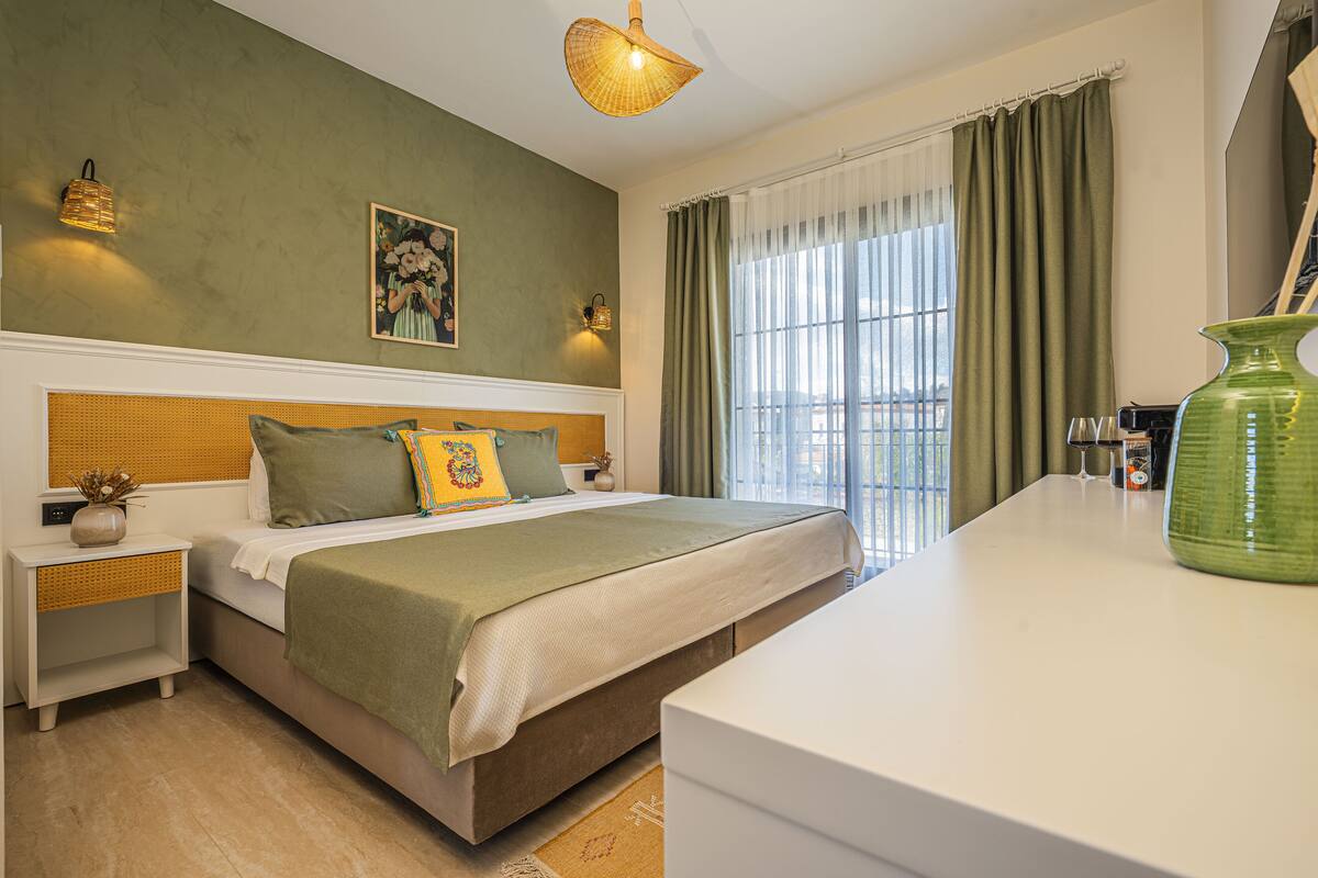 A bedroom with a large bed, two bedside tables, a nightstand, a green vase, and a window with curtains.