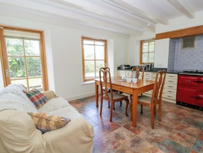 A cozy living room with a sofa, a dining table with chairs, a kitchen area with a red stove, and large windows. - Bryn Isaf (Tregaron)