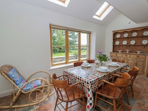 A dining area with a table set for four, surrounded by wicker chairs, a view of greenery outside, and a wooden cabinet with plates. - Bryn Isaf (Tregaron)