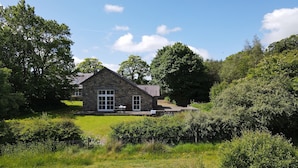A stone house with a sloped roof, large windows, and a patio area with a table and chairs. - Ty Cellin (Tregaron)