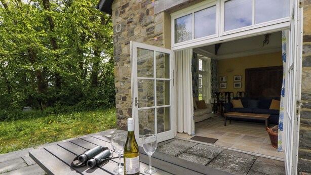 An outdoor patio with a stone wall, a wooden table with wine bottles and glasses, and a view into a room with a sofa and chairs. - Ty Cellin (Tregaron)