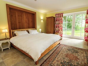 A bedroom with a large bed, wooden headboard, two bedside tables with lamps, a nightstand, a wooden wardrobe, and a view through a glass door. - Ty Cellin (Tregaron)