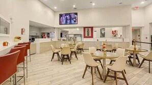 Ein moderner Essbereich mit mehreren Tischen und Stühlen, eine Küche im Hintergrund und ein an der Wand befestigtes Fernsehgerät.