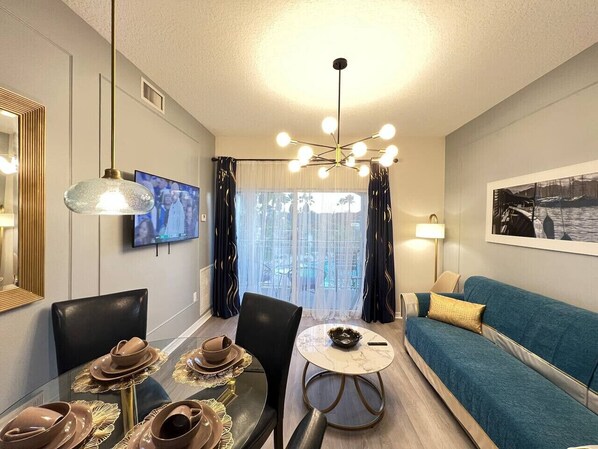 Een moderne woonkamer met een eethoek, een bank, een televisie en uitzicht op het groen door grote ramen.