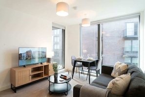 A modern living room with a flat-screen TV, a sofa, a dining table with chairs, and large windows. - Central Manchester 1BR Apartment with Balcony (Salford)