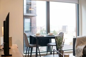 A dining area with a table set for two, a potted plant, and a view of the city through large windows. - Central Manchester 1BR Apartment with Balcony (Salford)