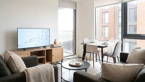 Una sala de estar moderna con televisor de pantalla plana, un comedor con mesa y sillas, y ventanas amplias que ofrecen vista a la ciudad.