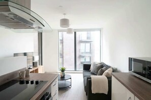 A modern kitchen with a stove, microwave, and dining area. There is a sofa and a small table with a plant. - Central Manchester 1BR Apartment with Balcony (Salford)