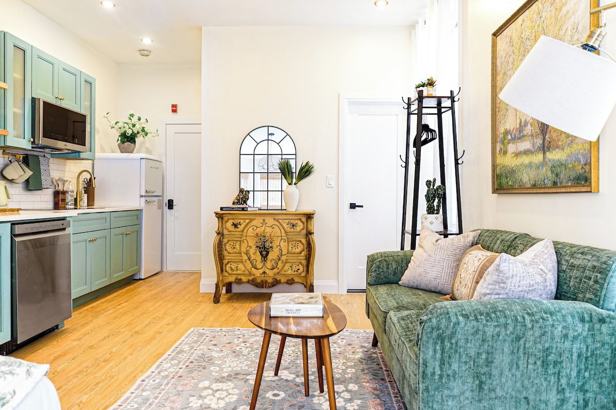 Un espace de vie compact comprenant un coin cuisine, un canapé vert, une commode décorative et un tableau accroché au mur.