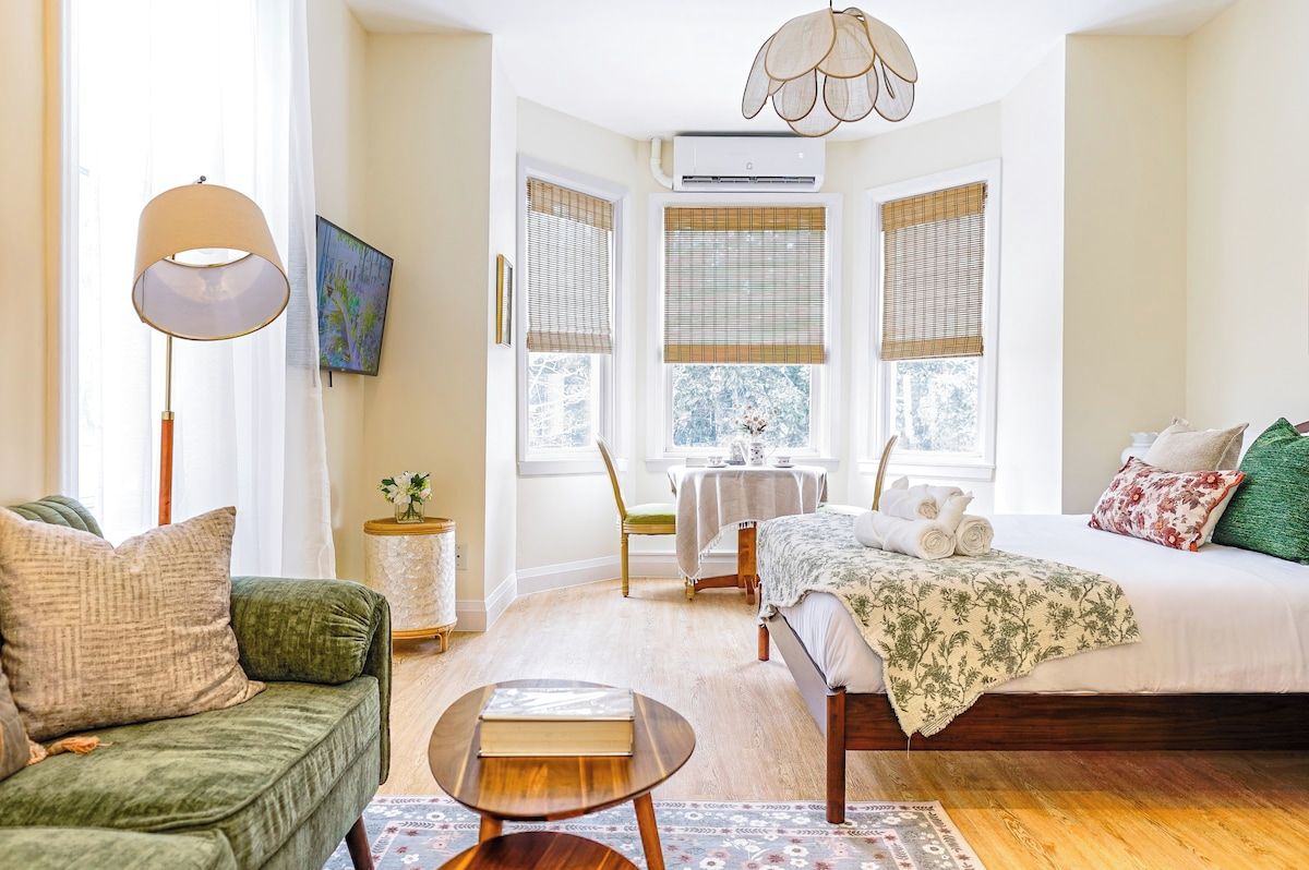 A bedroom with a bed, a bedside table, a chair, a lamp, and a window with blinds.