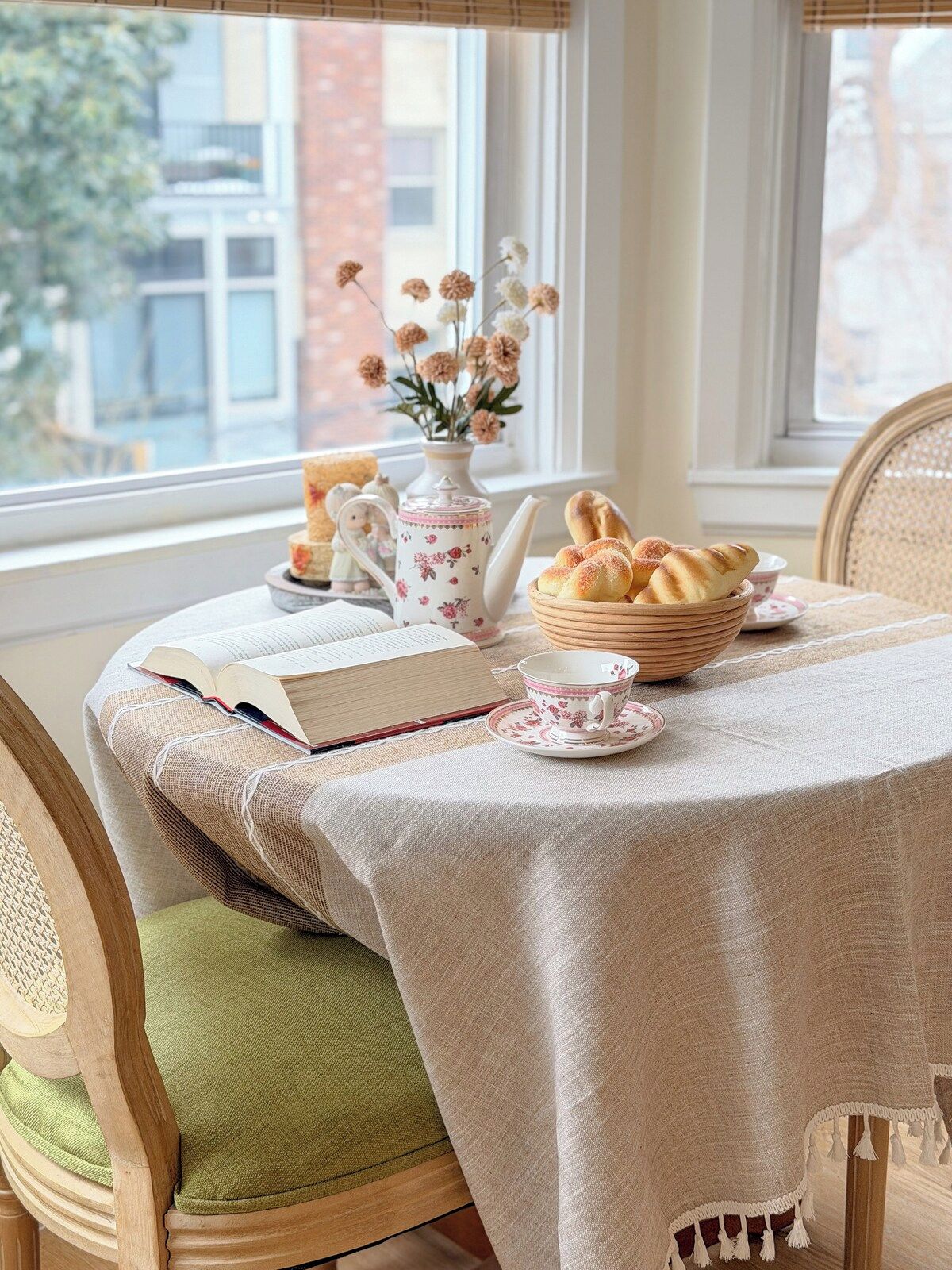 A dining table with a book, teapot, and pastries, set against a window with a view of buildings.