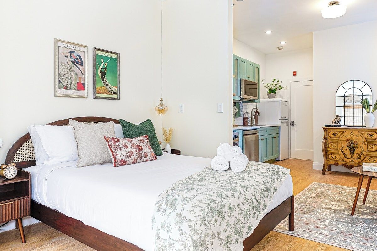 A bedroom with a bed, pillows, a bedside table, a clock, and a nightstand. There is a kitchen visible in the background.