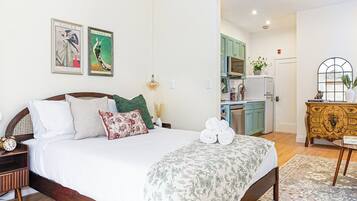 A bedroom with a bed, pillows, a bedside table, a clock, and a nightstand. There is a kitchen visible in the background.