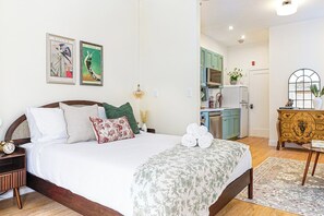 Un dormitorio con cama, almohadas, una mesita de noche, un reloj y una mesita auxiliar. Se ve una cocina al fondo.