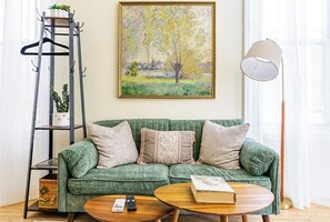 Un canapé vert avec trois coussins, une table basse en bois avec une télécommande et un livre, une lampe sur pied et un tableau représentant un arbre accroché au mur.