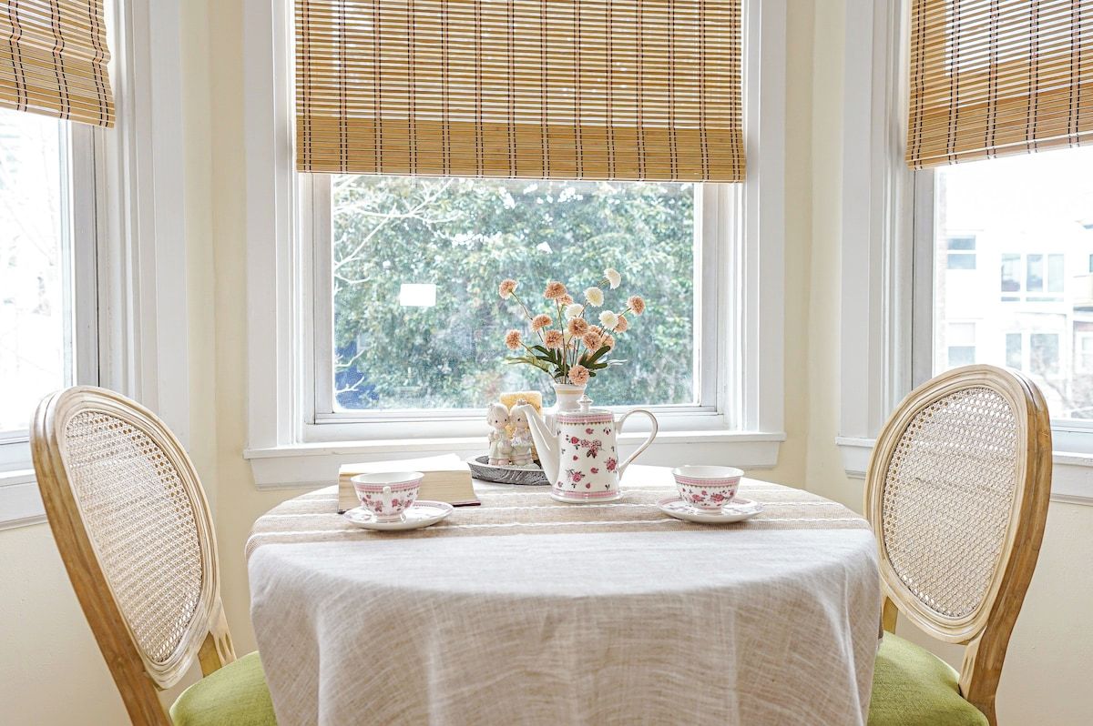 A dining area with a table set for two, a vase with flowers, and a teapot on the windowsill.