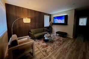 A modern living room with a wooden panel wall, a flat-screen TV mounted above a sofa, a coffee table, and a floor lamp. - Luxurious rustic home (Ferndale)