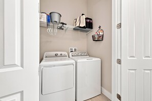 Une buanderie équipée de deux machines à laver et de deux sèche-linge à chargement frontal, tous deux de couleur blanche, d’une étagère en métal contenant des produits de lessive et d’un panier décoratif en métal accroché au mur.