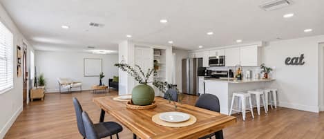 A spacious living area with a wooden dining table, modern kitchen, and a large window.