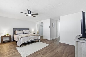 A bedroom with a bed, bedside tables, a ceiling fan, a television, and a hallway.