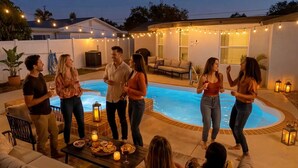 Une réunion conviviale au bord de la piscine, où les gens sont debout et discutent, entourés de meubles d’extérieur et de guirlandes lumineuses.