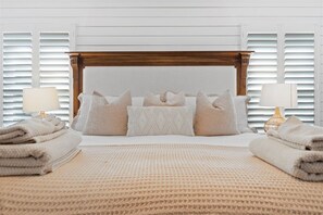 Een net opgemaakt bed met een beigekleurige deken met textuur, een houten hoofdbord en meerdere kussens. Aan beide zijden van het bed staan nachtlampjes en liggen gevouwen handdoeken.