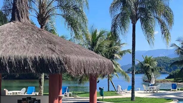 A pool area with a thatched-roof structure, lounge chairs, and palm trees. - Apartment at Costabella Resort in Angra dos Reis (Angra dos Reis)