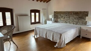 A bedroom with a large bed, a stone accent wall, a wooden ceiling, a radiator, and a wicker chair. - Apartment 'Casa La Corte in Capannori' with Private Pool, Wi-Fi & Air Conditioning (Capannori)