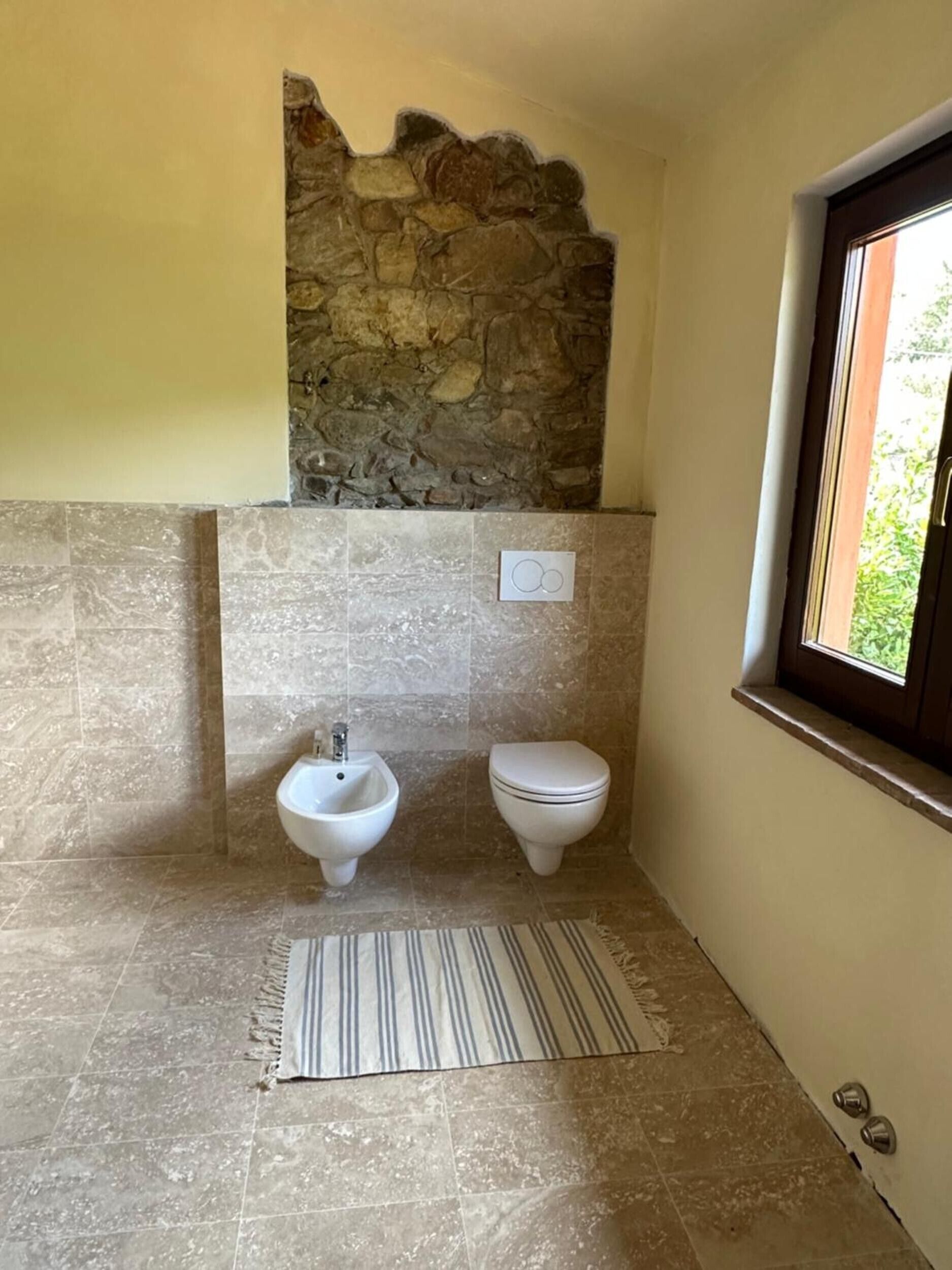 A bathroom with a stone wall, a bidet, a toilet, and a window.