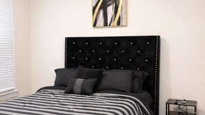 Quarto com cabeceira estofada preta, colcha listrada e pequena mesa de cabeceira com abajur.