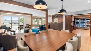 Un hall d’hôtel spacieux, doté d’une grande table en bois, de sièges confortables, de luminaires suspendus et offrant une vue sur le parking.